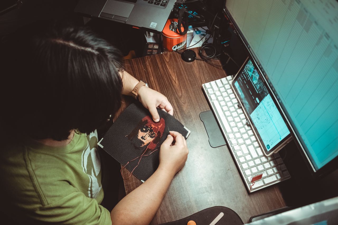 An adult stitching cross-stitch art at a desk with a computer on a wooden table.