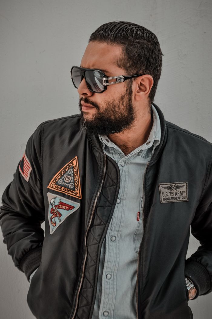 Portrait of a stylish man in a black jacket with patches and sunglasses posing confidently.
